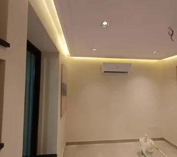 منظر داخلي لغرفة مكتب إدارية حديثة قيد التشطيب النهائي، يظهر سقف جبس مع إضاءة مخفية دافئة، سبوتلايت، وحدة تكييف سبليت، جدران مدهونة، ونوافذ سوداء، من تنفيذ مؤسسة تشطيبات في المنطقة الشرقية 